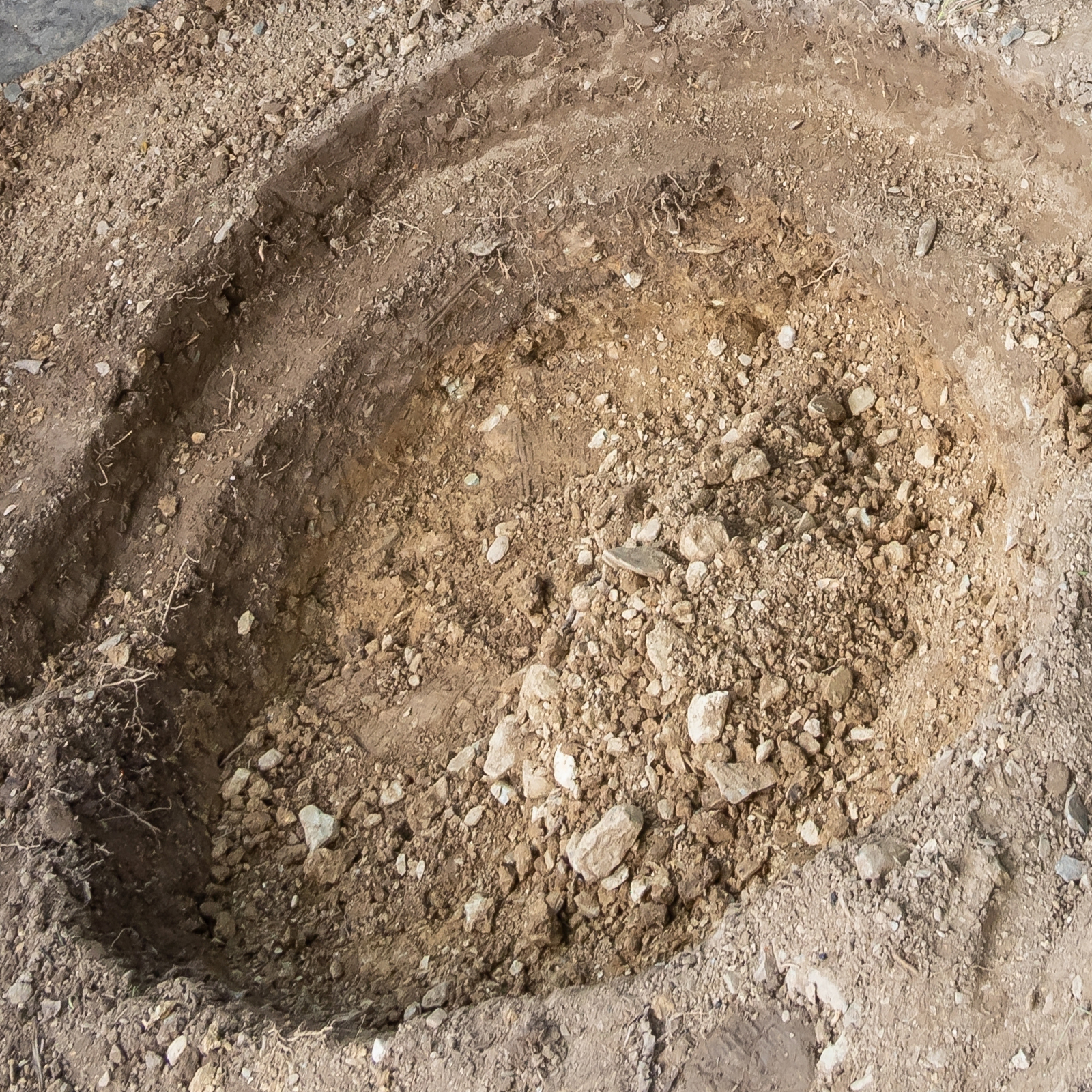 excavated dirt hole prepared for VEVOR pond liner installation, showing soil and rocky layers.