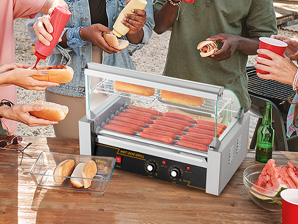 Draugi bauda bārbekjū pagalmā ar VEVOR hotdogu rullīti, hotdogiem, maizītēm un garšvielām uz galda.