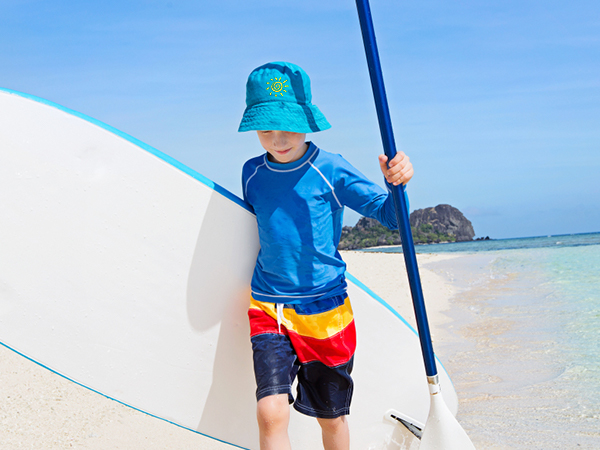 child on a tropical beach holding a paddleboard, wearing a sun hat, and colorful swim trunks.