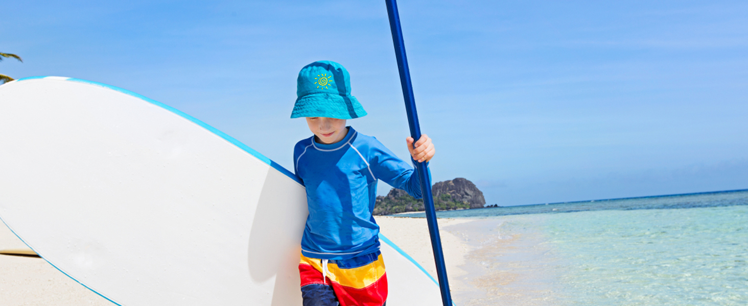 child on a tropical beach holding a paddleboard, wearing a sun hat, and colorful swim trunks.