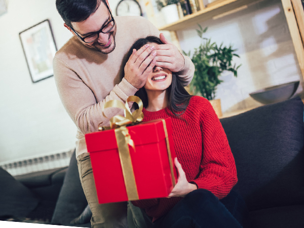 couple exchanging a red gift box with a golden ribbon in a cozy home setting.