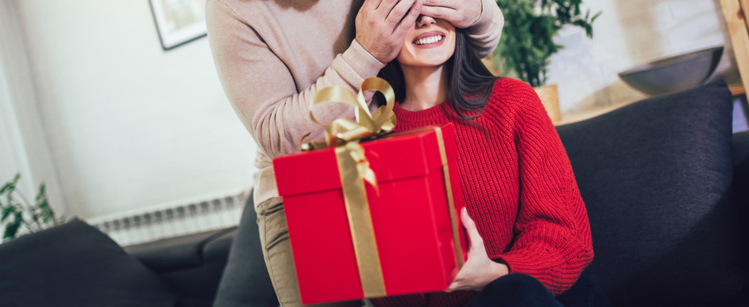 couple exchanging a red gift box with a golden ribbon in a cozy home setting.