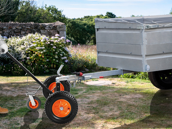 Cilvēks izmanto VEVOR piekabes ratiņus, lai manevrētu ar piekabi pa zāļainu reljefu netālu no akmens sienas ar ziediem.