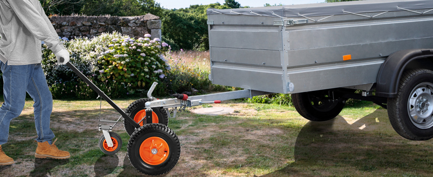 Cilvēks izmanto VEVOR piekabes ratiņus, lai manevrētu ar piekabi pa zāļainu reljefu netālu no akmens sienas ar ziediem.